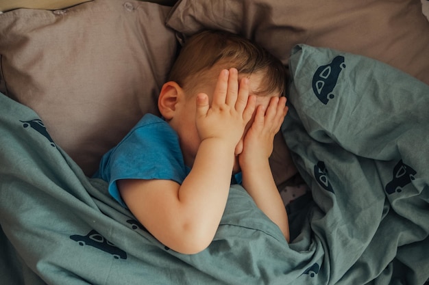 Un bébé en chemise bleue couvre son visage avec ses mains.