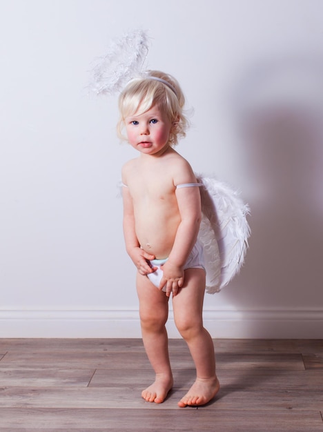 Le bébé en costume d'ange se tient sur un fond de mur blanc Un bébé blond nu est debout dans une couche