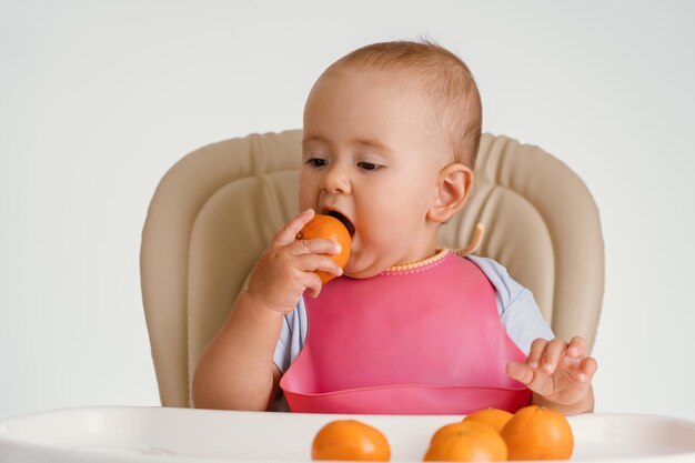 Un bébé dans un bavoir rose est assis sur une chaise essayant de manger une mandarine dans la peau