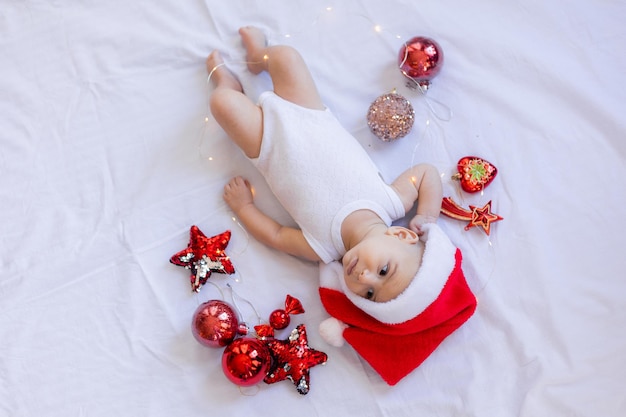 Bébé dans un body blanc et un bonnet de Noel est allongé sur le dos sur un drap blanc entouré de jouets d'arbre de Noël rouges. hiver, nouvel an. espace pour le texte. Photo de haute qualité