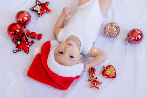 Bébé dans un body blanc et un bonnet de Noel est allongé sur le dos sur un drap blanc entouré de jouets d'arbre de Noël rouges. hiver, nouvel an. espace pour le texte. Photo de haute qualité