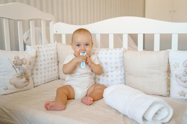 Un bébé dans un body blanc est assis dans un berceau et ronge une brosse à peigne