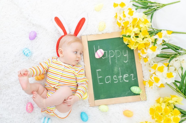 Un bébé dans un chapeau de lapin sur un lit avec des oeufs de Pâques et l'inscription Joyeuses Pâques