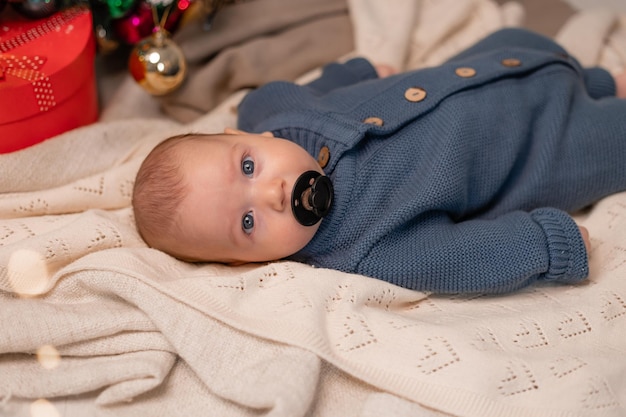 Bébé dans une combinaison tricotée bleue avec une tétine noire dans sa bouche est allongé sur une couverture légère sur le sol sous le sapin de Noël. premier Noël. vacances d'hiver, moments amusants. Photo de haute qualité