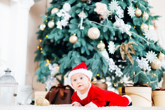 Un bébé dans un costume de père Noël est allongé sur le ventre près de l'arbre de Noël et des cadeaux