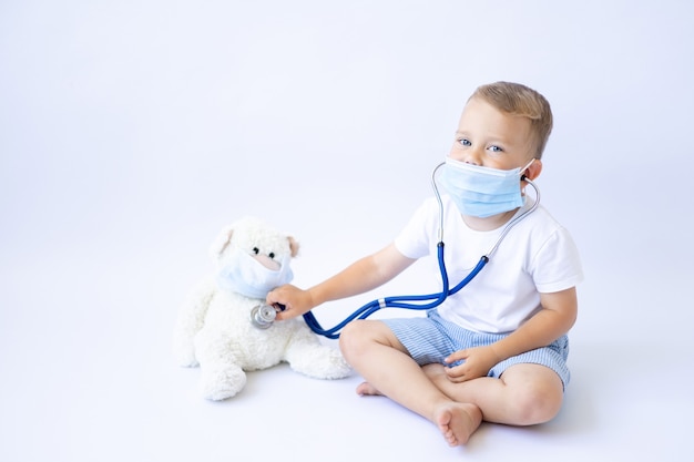 Un bébé dans un masque médical est assis avec un stéthoscope et traite une pandémie d'isolat de fond blanc en peluche