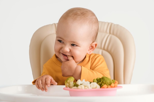 Bébé drôle sur une chaise d'alimentation tient ses doigts dans sa bouche. Une assiette de légumes bouillis devant lui