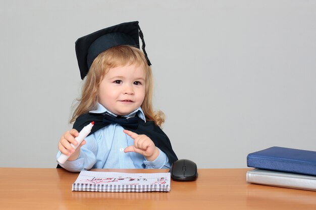 Bébé enfant dans le manteau académique et cap assis au bureau en bois de l'école tenant la marque