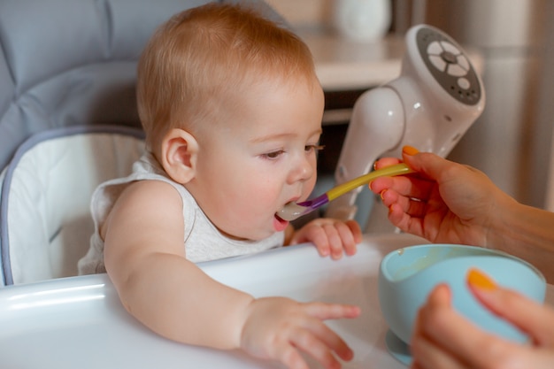 Le bébé est assis sur une chaise haute dans la cuisine, la main de sa mère nourrit la bouillie