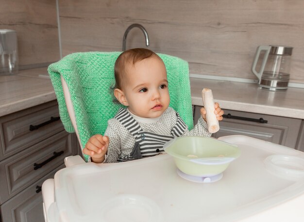 Bébé est assis dans une chaise haute dans la cuisine et mange une banane.