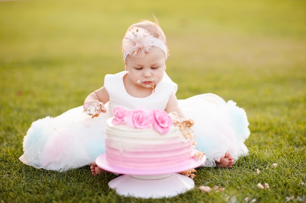 Bébé fille de 1 ans porter une robe de princesse en train de manger un gâteau au fromage à la crème d'anniversaire assis sur l'herbe verte à l'extérieur Célébration