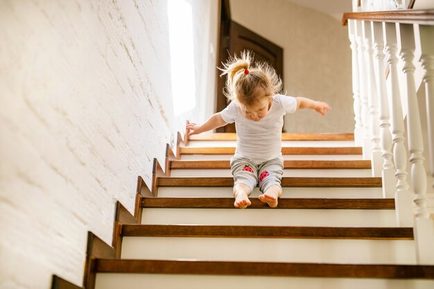 Bébé fille blonde en t-shirt blanc au bas des escaliers à l'intérieur, regardant la caméra et souriant.