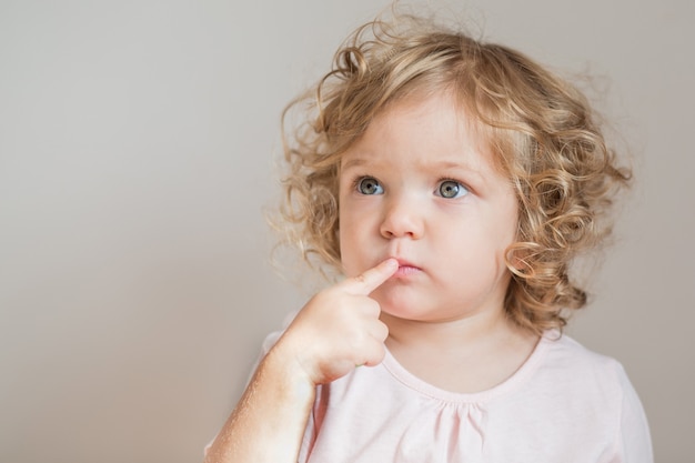 Bébé fille bouclée émotionnelle avec le doigt près de son visage sur un mur gris clair