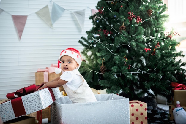 Un bébé garçon africain de 9 mois debout dans une boîte-cadeau près d'un sapin de Noël et de nombreux coffrets cadeaux