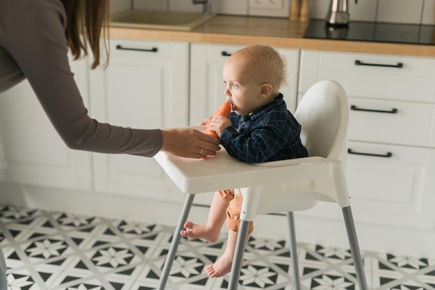 Bébé garçon assis dans une chaise bébé manger des carottes sur fond de cuisine espace de copie concept d'alimentation de bébé