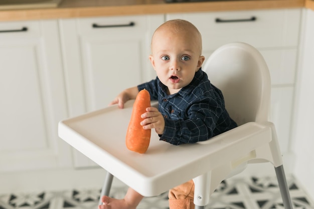 Bébé garçon assis dans une chaise bébé manger des carottes sur fond de cuisine espace de copie concept d'alimentation de bébé