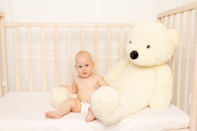 Bébé garçon assis dans des couches dans un berceau avec un ours en peluche