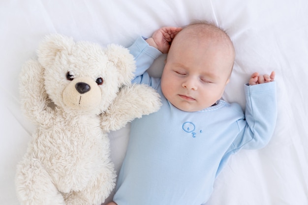 Bébé garçon dort sur le lit allongé sur le dos avec un ours en peluche en pyjama bleu