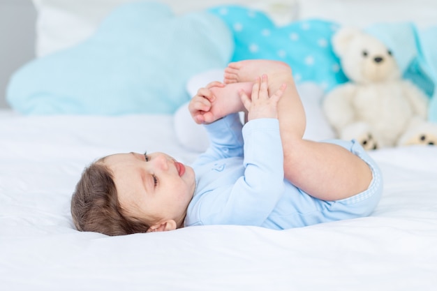 Bébé garçon sur le lit dans un body bleu jouant avec ses pieds, heureux petit bébé en bonne santé