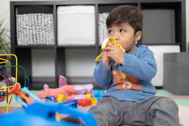 Bébé garçon mexicain de deux ans mangeant de la banane dans une chambre en désordre