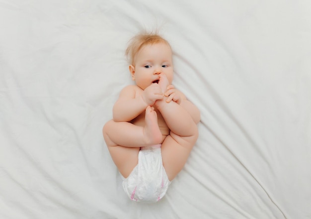 Un bébé heureux et joyeux en couches est allongé sur un lit blanc et tient sa jambe Lieu de photo de haute qualité pour le texte