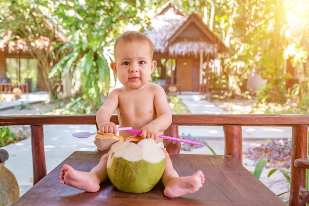 Bébé heureux en vacances tropicales Mange et boit une jeune noix de coco verte S'assoit sur le sol