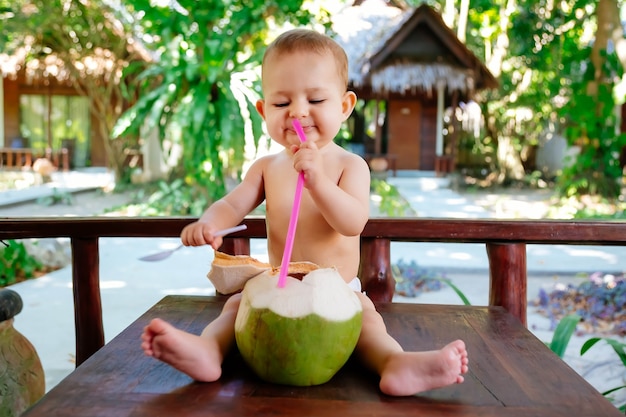 Bébé heureux en vacances tropicales Mange et boit une jeune noix de coco verte S'assoit sur le sol