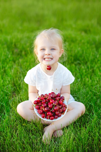 Un bébé joyeux assis sur l&#39;herbe verte avec une assiette de cerises et tenant une baie dans ses dents.