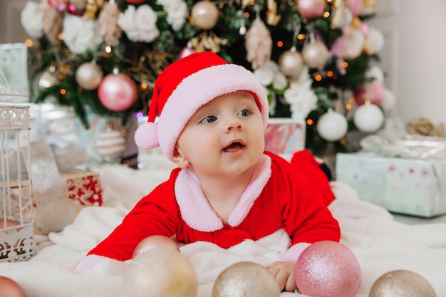 Un Bébé Mignon De 6 Mois Dans Un Costume De Père Noël Se Trouve Sur Une Couverture Près De L'arbre De Noël. Noël 2022