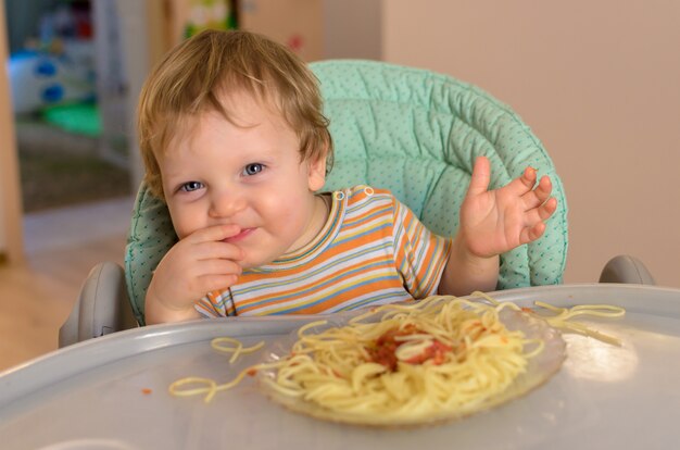 Un bébé mignon d'un an mange des spaghettis dans une chaise pour enfants.