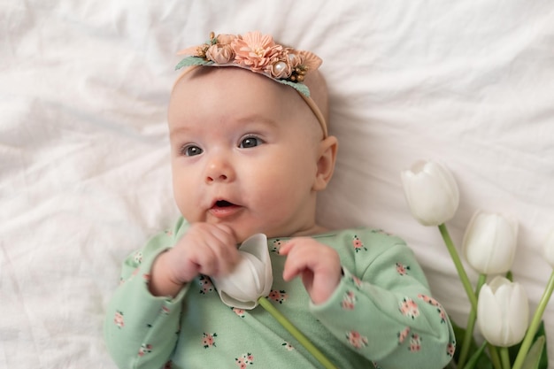 Bébé mignon avec un bandeau floral et un body en coton vert à la maison sur un lit blanc avec des tulipes. Spr