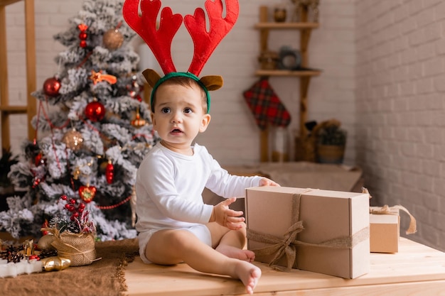 Un bébé mignon dans un body blanc et des cornes de renne est assis avec des cadeaux dans la cuisine à la maison pour Noël l'enfant regarde des cadeaux pour la nouvelle année