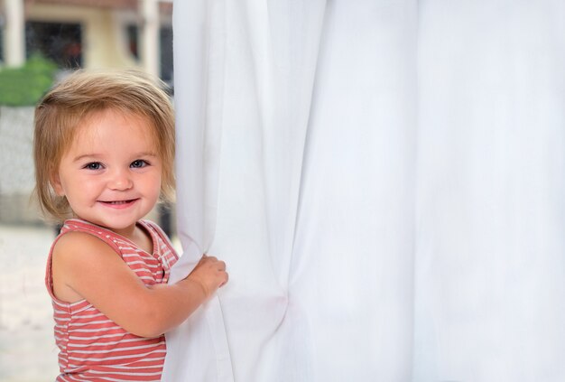 Un bébé mignon joue avec des rideaux. Portrait de l'enfant près de la fenêtre