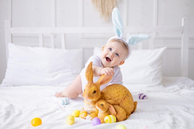 bébé mignon avec des oreilles de lapin et des œufs colorés sur un lit blanc à la maison jouant petit bébé blond avec un lapin bon concept de Pâques