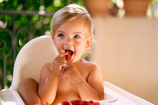 Bébé mignon s'asseyant sur une chaise haute et mangeant des fraises