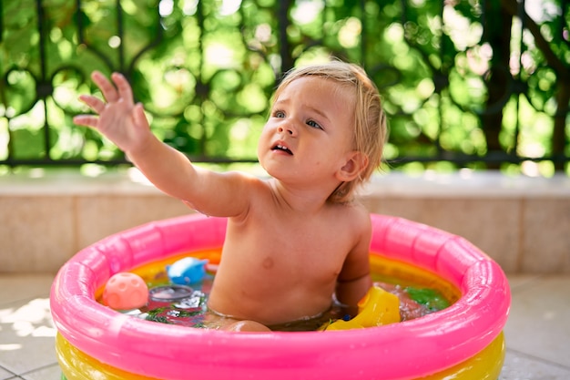 Le bébé mignon s'assied dans une petite piscine gonflable avec des jouets et tire sa main vers le haut