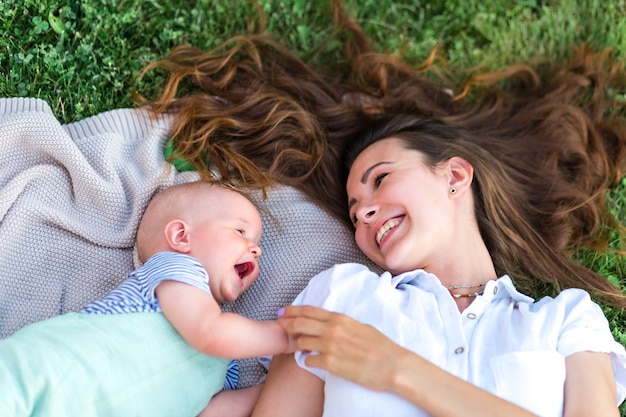 Le bébé mignon et sa maman se regardent et rient, ils sont allongés sur le dos sur l'herbe de la pelouse et bl...