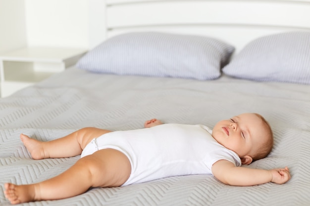 Bébé paisible allongé sur un lit tout en dormant dans un lit moelleux sur une couverture grise, un nourrisson portant un body blanc dort seul à l'intérieur, enfance.