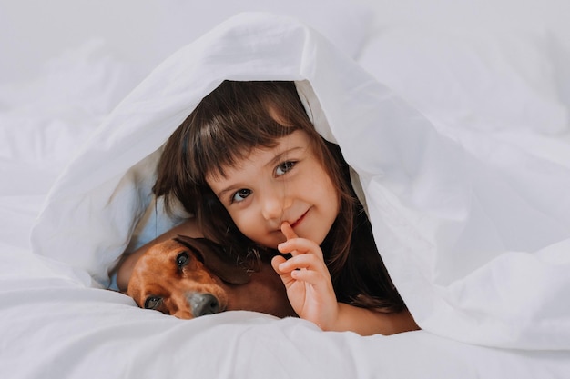 bébé petite fille embrasse un chien teckel à la maison dans son lit. linge de lit blanc. amour pour les animaux de compagnie