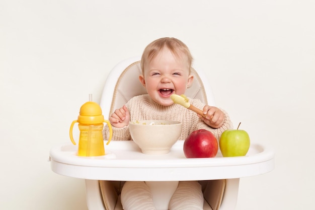 Bébé portant un pull tricoté assis dans une chaise haute et a faim en tenant une cuillère et en mangeant de la purée ou du porridge en dégustant des pommes et de l'eau dans une bouteille jaune posant isolé sur fond blanc