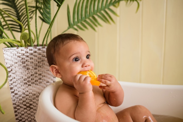Bébé se baigne dans un bain et mange une orange sur un mur en bois