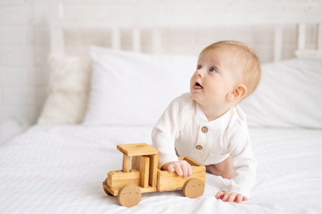 Un bébé souriant de 6 mois, un garçon blond est assis sur un grand lit dans une chambre lumineuse et joue avec une petite voiture en bois dans un body en coton le concept de produits pour enfants