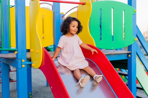 Photo bébé swarty aux cheveux bouclés en robe rose pâle joue sur une aire de jeux de rue et descend la colline