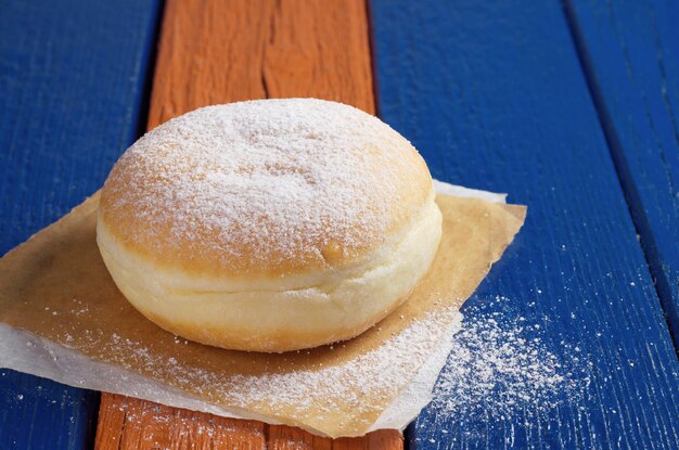 Le beignet berlinois avec de la confiture et du sucre en poudre sur papier est situé sur une table en bois colorée en gros plan