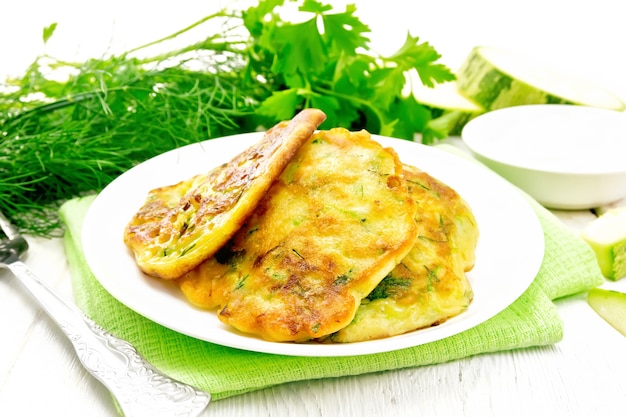 Beignets de courgettes, aneth et persil dans une assiette sur une serviette, crème sure dans une assiette sur fond de planche de bois