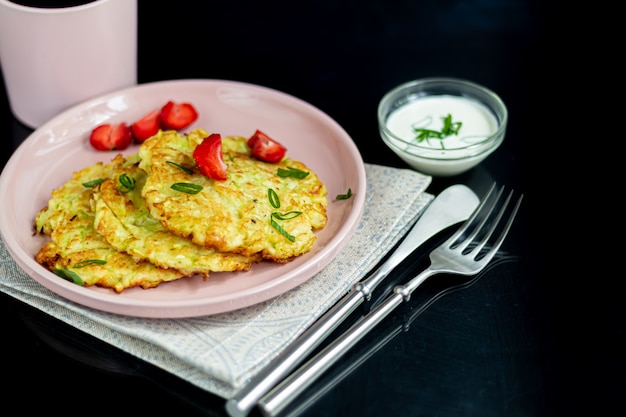 Beignets de courgettes, beignets végétariens de courgettes, servis avec des herbes fraîches et de la crème sure. Garni de fraises et d'oignons verts. Sur une table sombre.