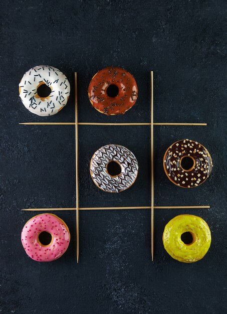 Beignets multicolores avec glaçage et arrose sur fond noir jeu de tic-tac-toe.