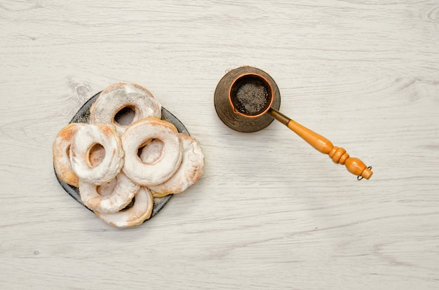 Beignets saupoudrés de sucre en poudre, café frais en cezve, fond en bois clair. Vue de dessus