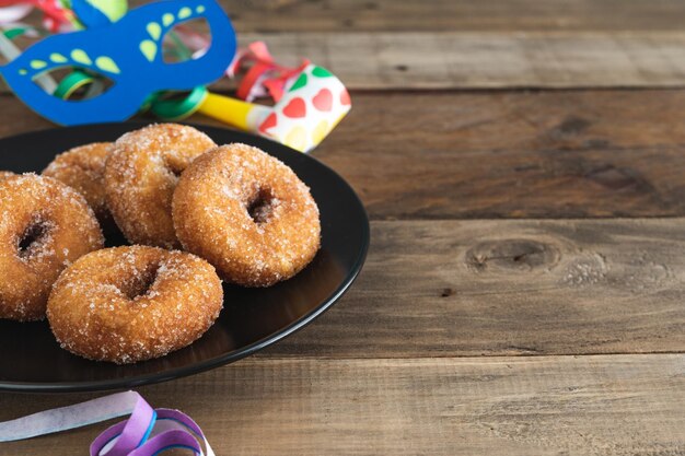 Photo des beignets sucrés sur une assiette noire sur un fond en bois avec des décorations typiques du carnaval.