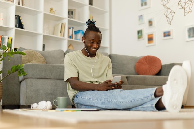 Un bel adolescent afro-américain envoie des SMS assis par terre à la maison sur un smartphone.